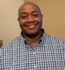 Smiling person wearing a colorful plaid shirt, standing against a neutral background. Headshot.