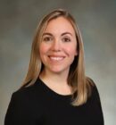 Headshot of a person with long, straight, light brown hair, smiling and wearing a black top, against a neutral background.