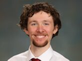 Headshot of a person with curly hair, wearing a white dress shirt and red tie, smiling against a blurred background.