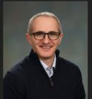 Headshot of a person wearing glasses and a dark zip-up shirt against a blurred background, smiling at the camera.