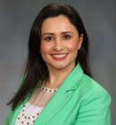Headshot of a person wearing a green blazer and white top, smiling, with a blurred background.